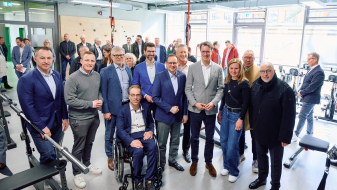 Hendrik Wüst umgeben von mehreren Personen, davon einer im Rollstuhl, in einer Halle mit Fenstern im Hintergrund