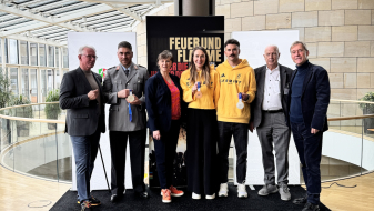 Sieben Personen auf einem schwarzen Podest im Landtag NRW mit vielen Fenstern im Hintergrund