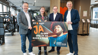 Andrea Milz mit drei Männern, im Hintergrund ist ein Fitnessstudio zu sehen, sie halten ein Banner "Fitx sagt Ja zu Spielen an Rhein & Ruhr"