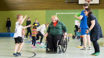 Ältere Frau in Sportkleidung im Rollstuhl ist umgeben von jüngeren Sportlern in eienr Sporthalle