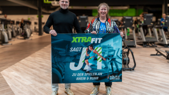 Andrea Milz neben einem Mann in einem Fitnessstudio, beide halten ein Banner zur KRR-Kampagne