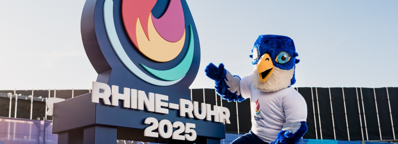 Das Maskottchen der Spiele vor dem Logo der Spiele